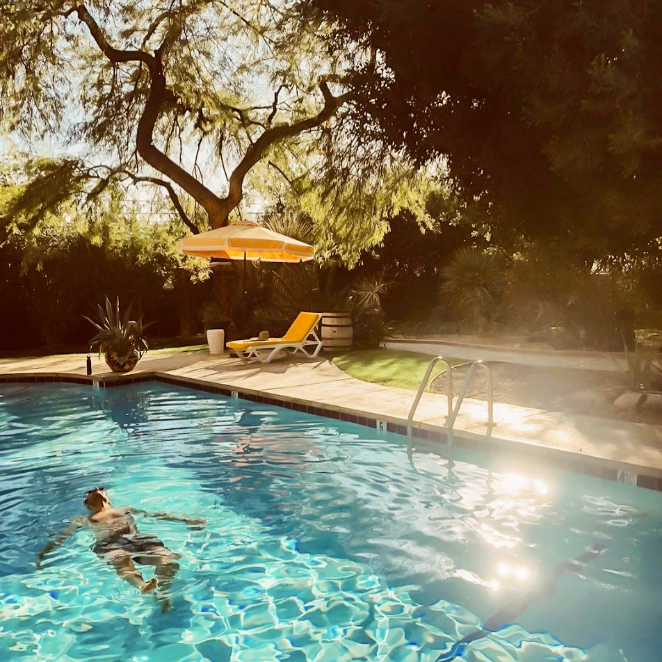 Guest swimming in a sunlit mineral pool beside a yellow umbrella and lounge chairs at a Miracle Hill spa resort in Desert Hot Springs, California