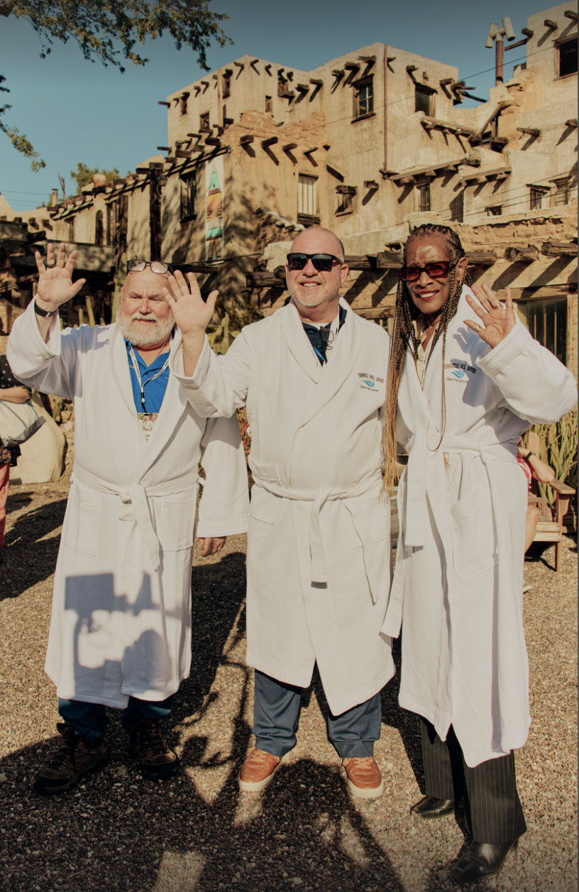 Three smiling guests in white Miracle Hill Spas robes waving at a Desert Hot Springs spa event